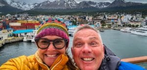 Smiling couple in winter jackets with Ushuaia's port and snow-capped mountains in the background.