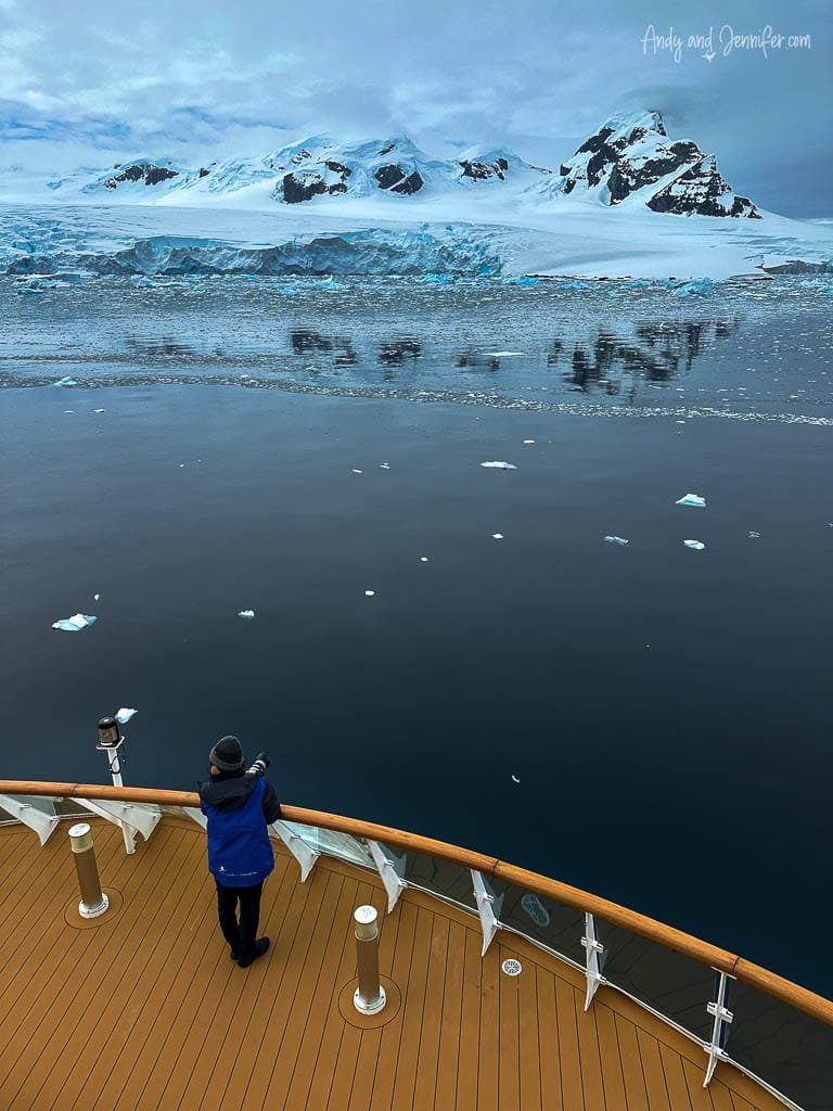 Person standing on a ship's deck, gazing at a stunning Antarctic scene with calm waters, scattered ice, and a backdrop of snow-covered mountains and glaciers.