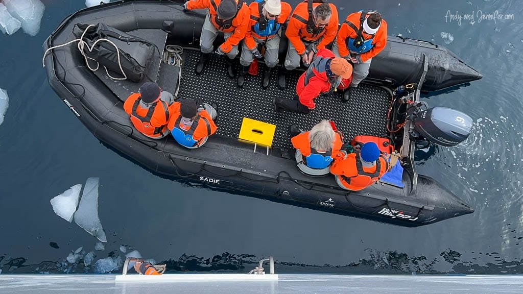 Group of people in orange life jackets seated in a black inflatable boat on icy water.