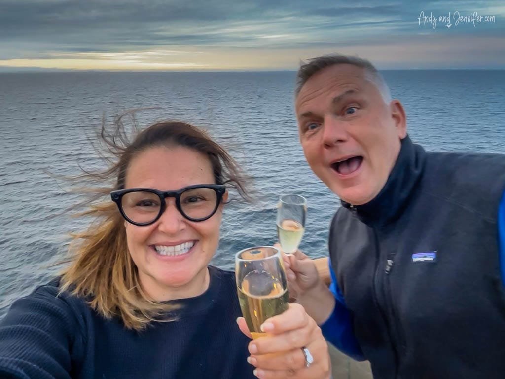 A smiling couple holds champagne flutes while standing on the deck of a cruise ship. The ocean stretches out behind them under a cloudy sky during sunset. The woman wears glasses and a black sweater, and her windblown hair adds a lively touch. The man, in a black vest, has an excited expression.