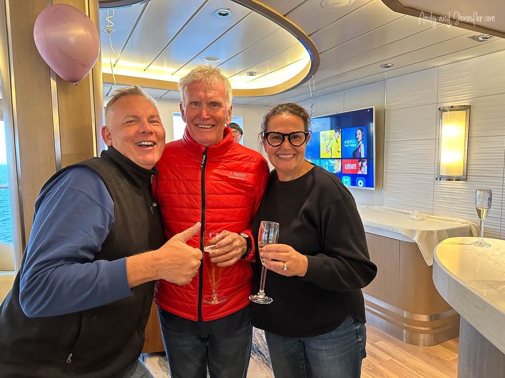 Three smiling people celebrate together inside a cruise ship, holding champagne flutes. One person, wearing a black vest, gives a thumbs-up, while the second, in a bright red jacket, stands between them. The third person, wearing black glasses and a black sweater, also holds a glass. A pink balloon hangs in the background, contributing to the festive atmosphere.