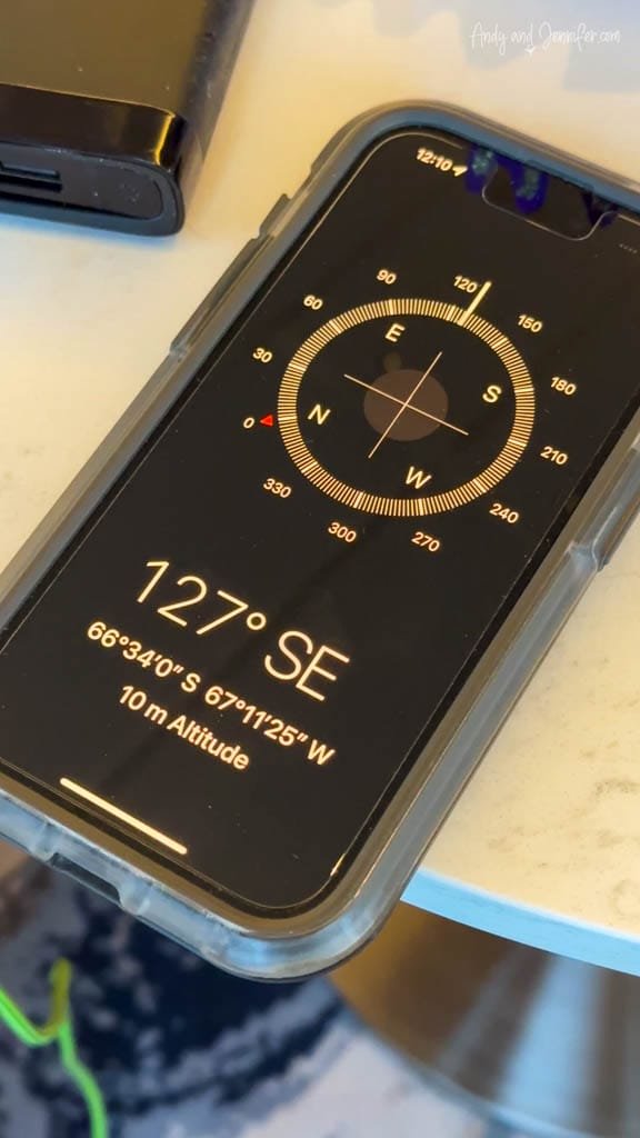 A smartphone with a compass app is displayed, showing the direction 127° SE. The coordinates read "66°34'0" S, 67°11'25" W" and the altitude is listed as "10 m." The phone is in a protective case, placed on a table alongside another device. 