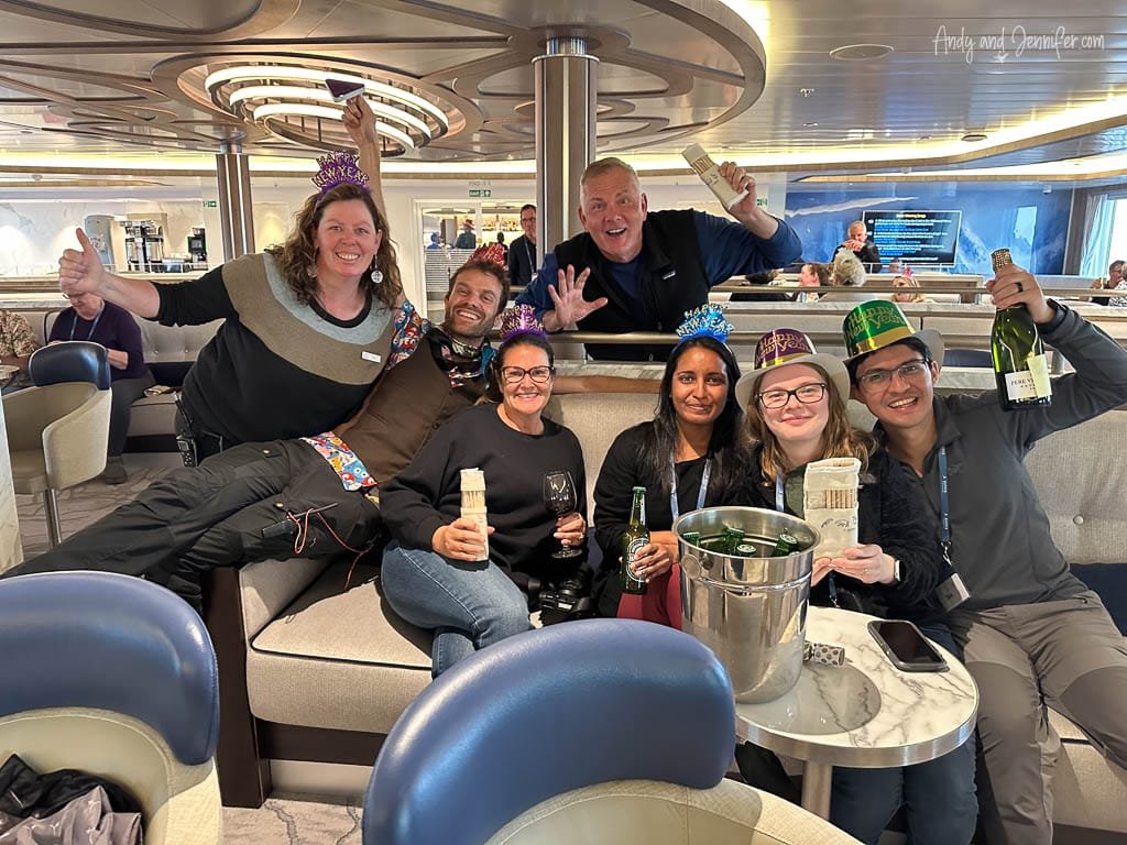 A group of seven people in festive New Year's hats celebrate onboard a ship, holding bottles of champagne and drinks. They are seated in a comfortable lounge area, smiling and posing joyfully for the camera. Party decorations and happy energy fill the space as they ring in the New Year together.