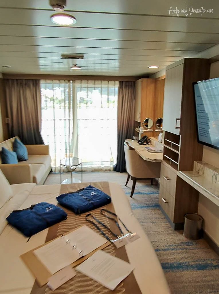  Modern cruise ship cabin with a bed, desk, and sliding glass doors leading to a balcony. Blue jackets and paperwork are neatly laid out on the bed.