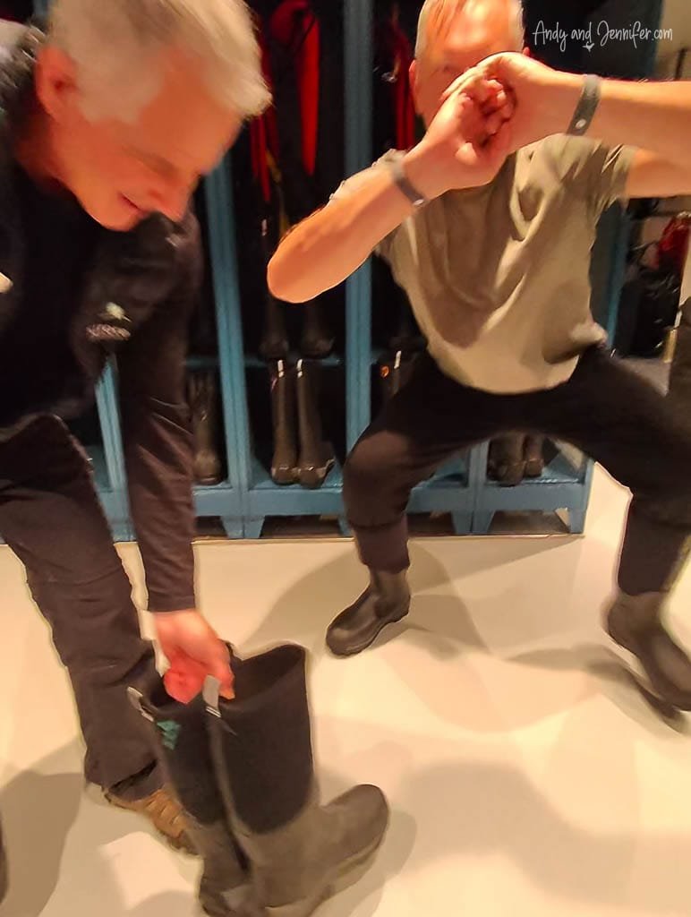Two men trying on sturdy black expedition boots inside a gear room, preparing for an Antarctic excursion.