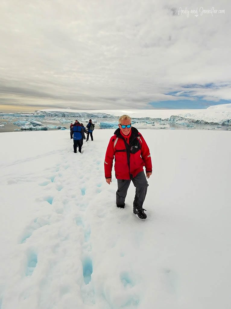 Man in red jacket and sunglasses walking on snow with group of people in icy landscape.