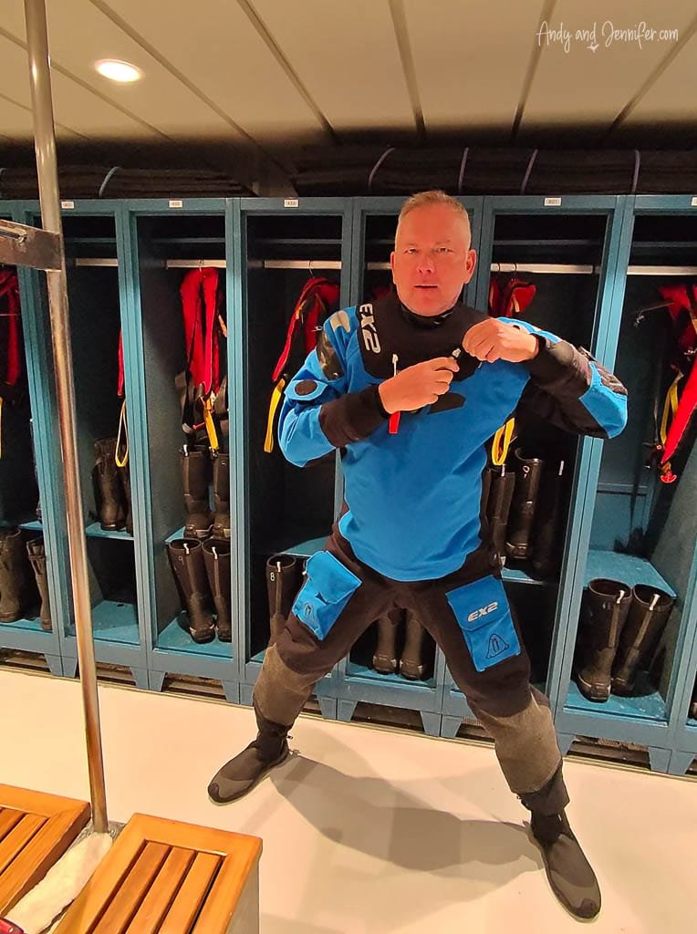Man in a blue drysuit preparing for an Antarctic expedition inside a gear room, with lockers and boots in the background.