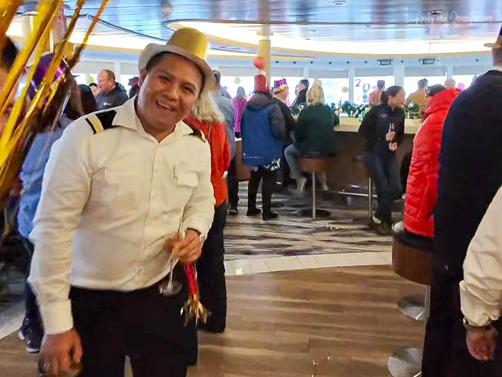 A smiling man in a white uniform and a gold party hat celebrates New Year's Eve on a cruise ship. He holds a drink and party noisemakers as people in festive attire gather around the bar in the background, enjoying the lively atmosphere.