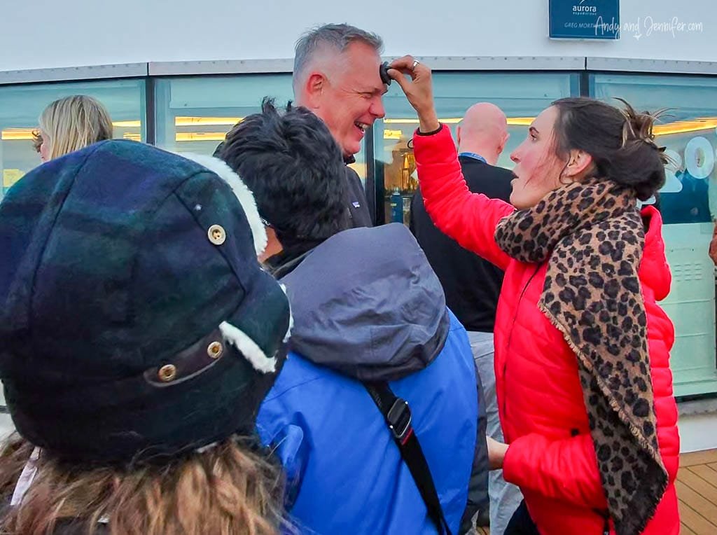 A woman in a red jacket and leopard-print scarf draws on a man's forehead with a black marker while he laughs. They are surrounded by people dressed warmly in winter jackets and hats, likely on the deck of a ship during a fun group activity. The atmosphere is lighthearted and social.