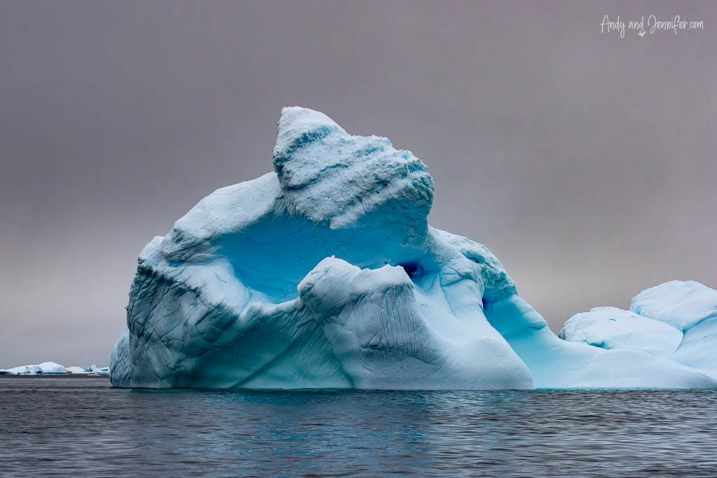 A large, jagged iceberg floats in Antarctic waters, its deep blue hues highlighted under a gray, overcast sky.
