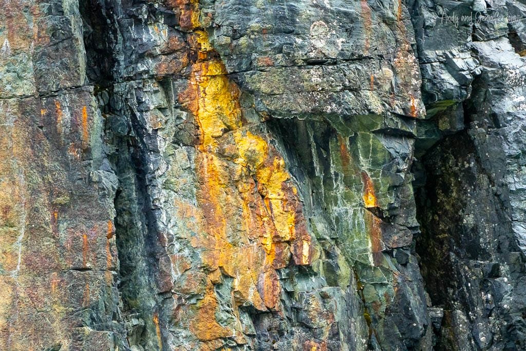 Close-up of a rugged cliff face featuring dark gray and green rock with streaks of bright yellow and orange mineral deposits. 