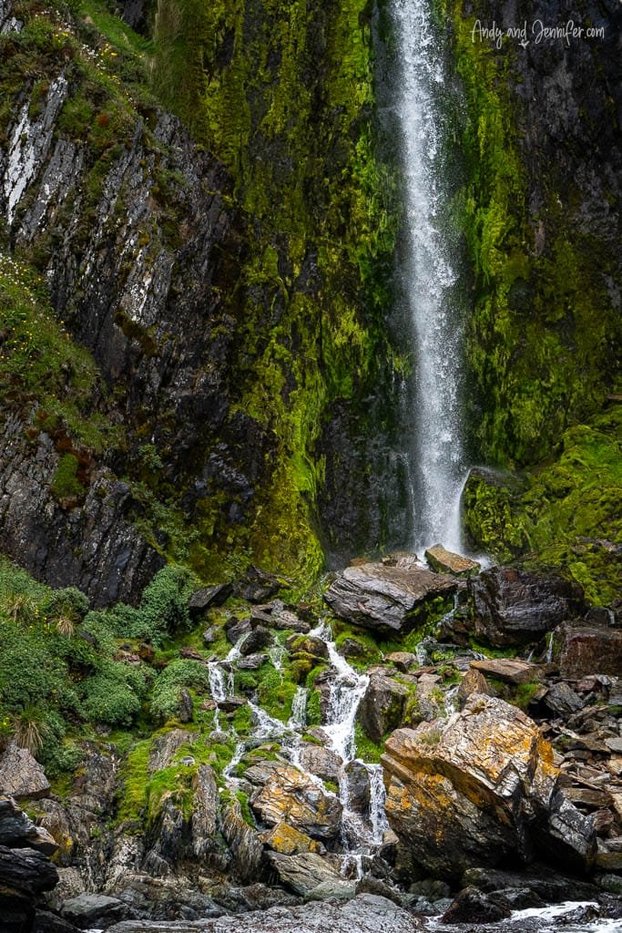 A tall waterfall cascades down moss-covered rocks, with smaller streams flowing over boulders at the base.