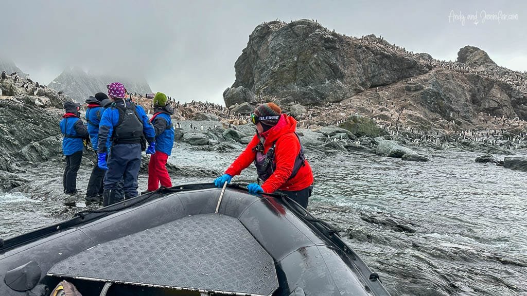 Group of people disembarking from a small boat onto rocky shore, surrounded by a large penguin colony under foggy skies.