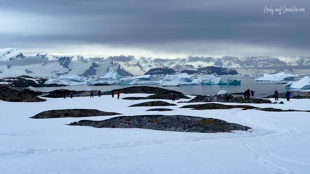 People exploring a snowy Antarctic landscape with icebergs and mountains under a cloudy sky. 