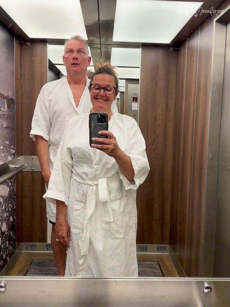 A man and woman, both wearing white bathrobes, pose for a humorous selfie in an elevator. 