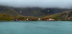 A distant view of an abandoned whaling station along the shoreline, with rusted buildings and tanks, nestled beneath fog-covered mountains.