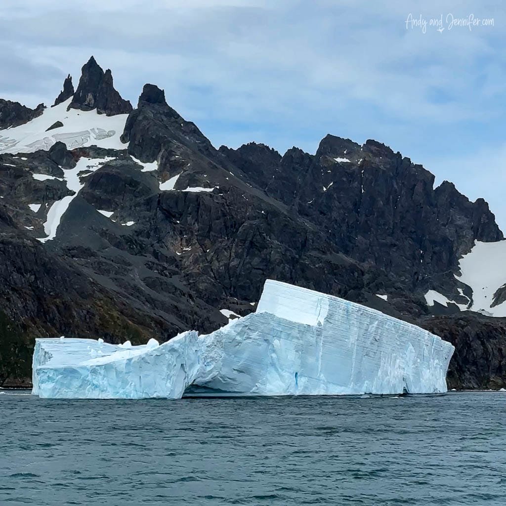 A massive iceberg with jagged edges floats near rugged, snow-dusted mountains.
