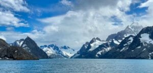 Snow-capped mountains rise sharply above a blue fjord, partially covered in clouds under a bright sky.