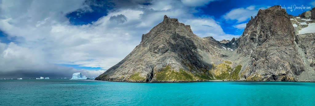 Panoramic view of towering rocky mountains meeting the vibrant turquoise sea, with distant icebergs floating under a partly cloudy sky.