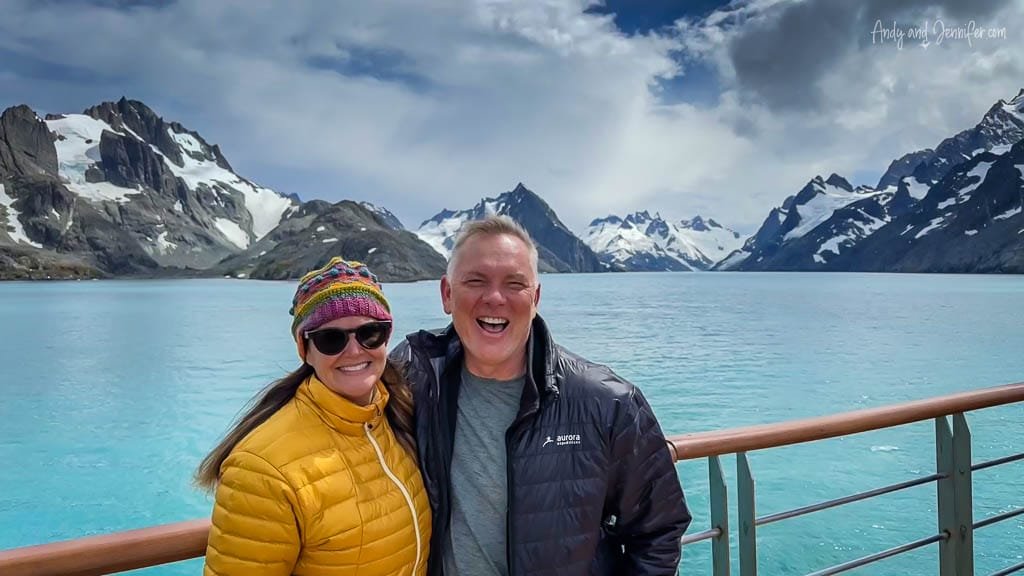 A smiling couple poses on a ship's deck with turquoise water, snowy mountains, and a dramatic sky in the background.