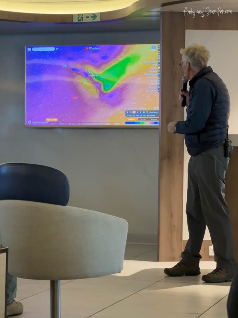 A man gives a presentation in a modern room, pointing to a colorful weather map displayed on a screen.