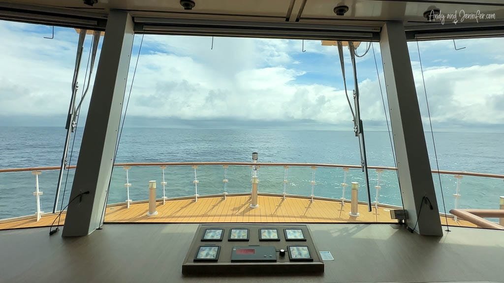 View from a ship's bridge looking out over the ocean through large windows, with control panels in the foreground and a wooden deck below.