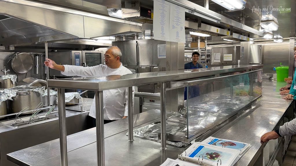 Chef giving a tour in a commercial ship kitchen, gesturing toward industrial cooking equipment while guests observe from behind a serving counter.