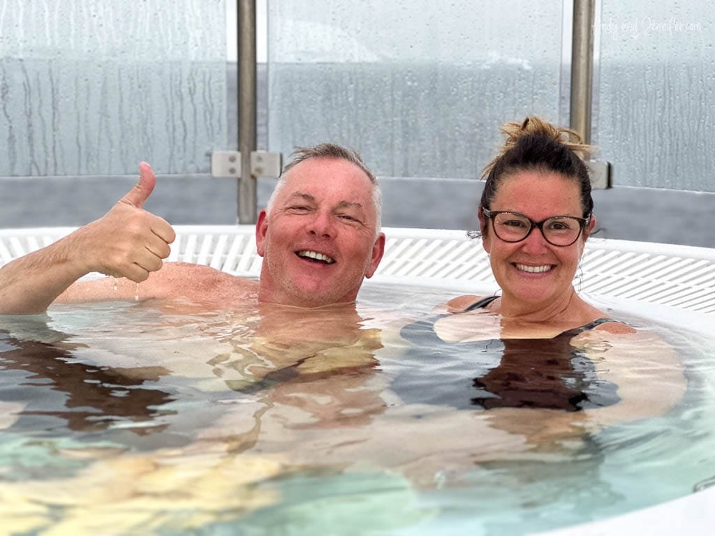 A man and woman relax in a hot tub, smiling at the camera. The man gives a thumbs-up gesture, and the woman wears glasses, both enjoying the warm water. The background shows a foggy or misty glass enclosure.