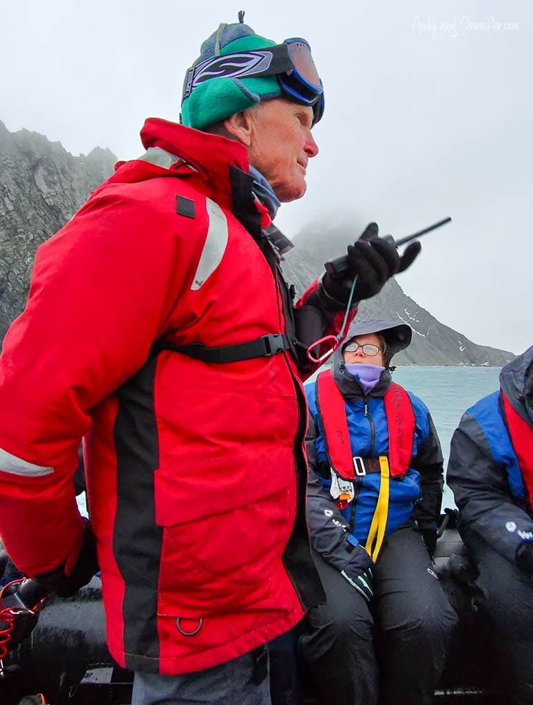 Person in a red jacket standing on a boat holding a radio, with others seated in blue jackets, surrounded by foggy mountains and the ocean.