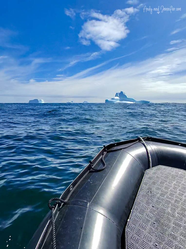 A Zodiac boat floats on calm blue water under a partly cloudy sky, with distant icebergs visible on the horizon.