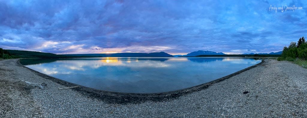 Panoramic view of a calm lake at sunset, with mountains in the distance and a pebble shoreline in the foreground