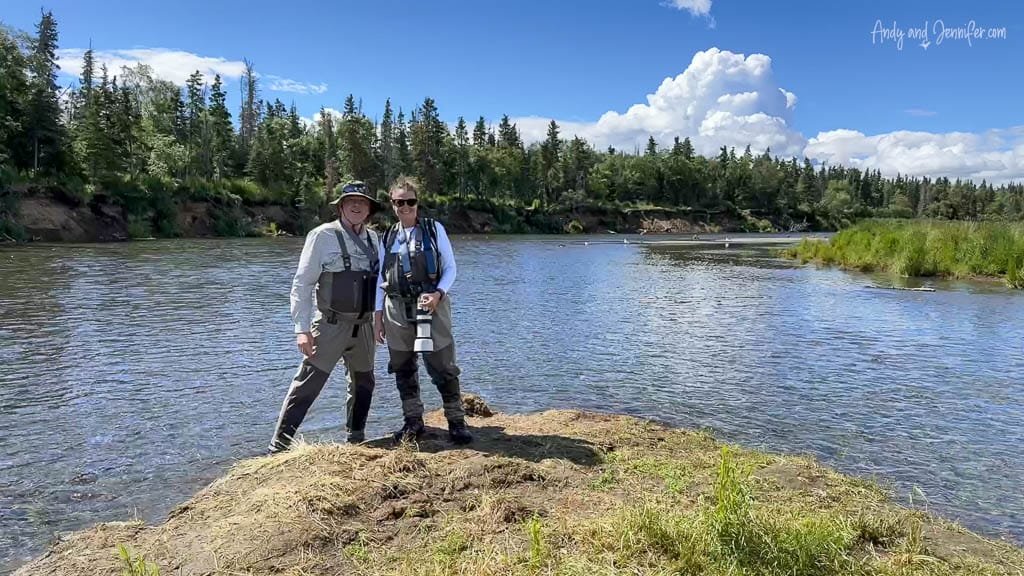 Two people in fishing gear posing by a river under a clear blue sky