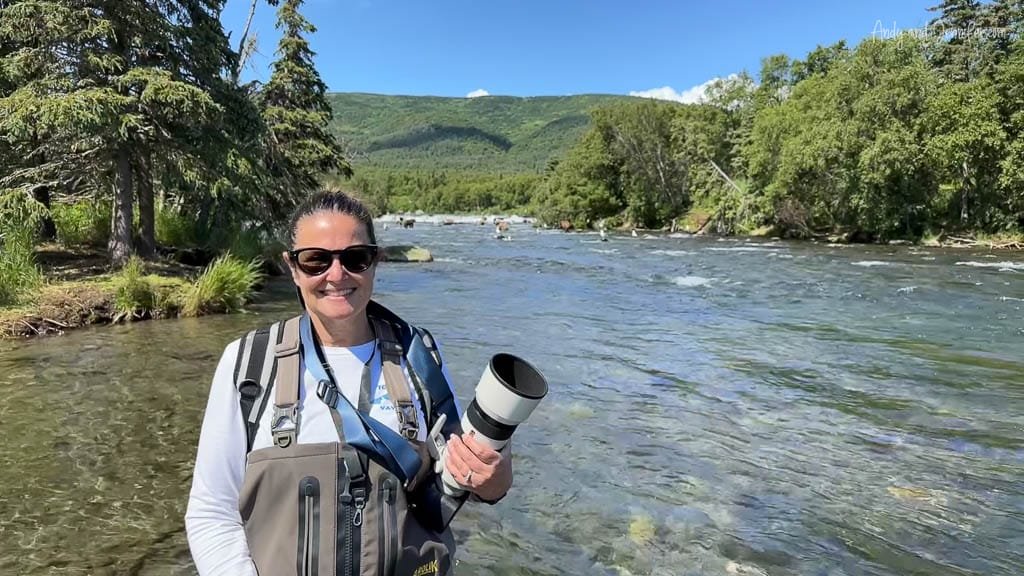 Smiling person with a camera standing in a river, surrounded by lush trees and mountains