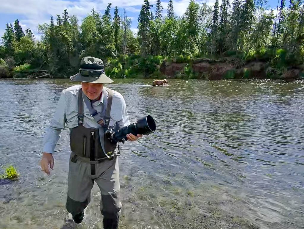 Photographer wading through a river with a camera, a bear visible in the background