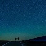 A couple holding hands, silhouetted against a vast, starry night sky on a deserted road, with a faint mountain range in the distance.