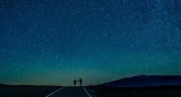 A couple holding hands, silhouetted against a vast, starry night sky on a deserted road, with a faint mountain range in the distance.