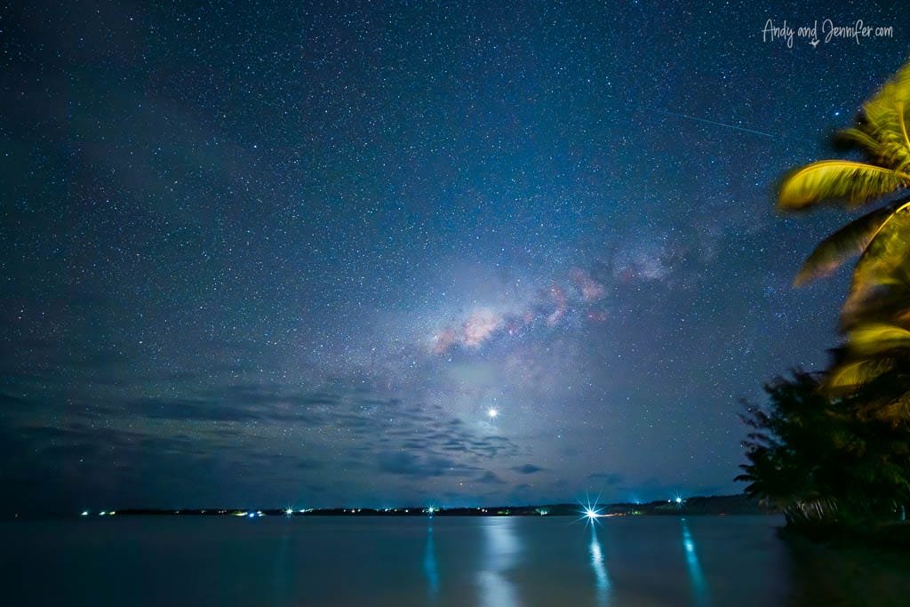  The Milky Way stretches across the night sky over Aitutaki's lagoon, framed by palm trees and shimmering water.
