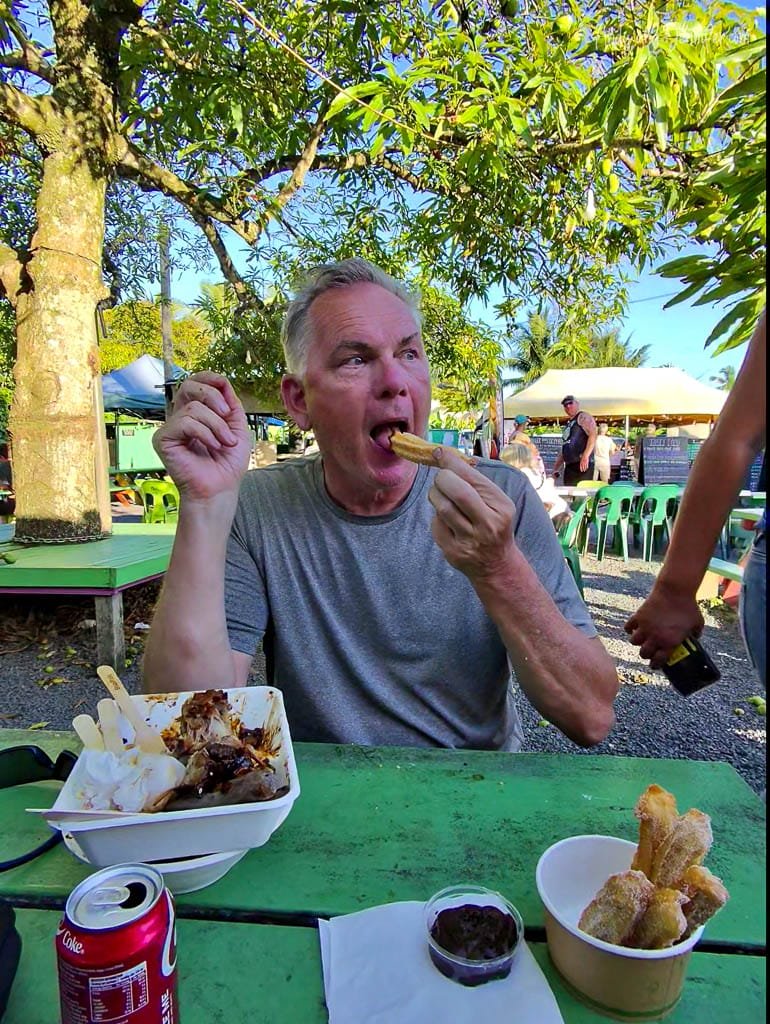 Man enjoying outdoor meal with churros and dessert at a market.