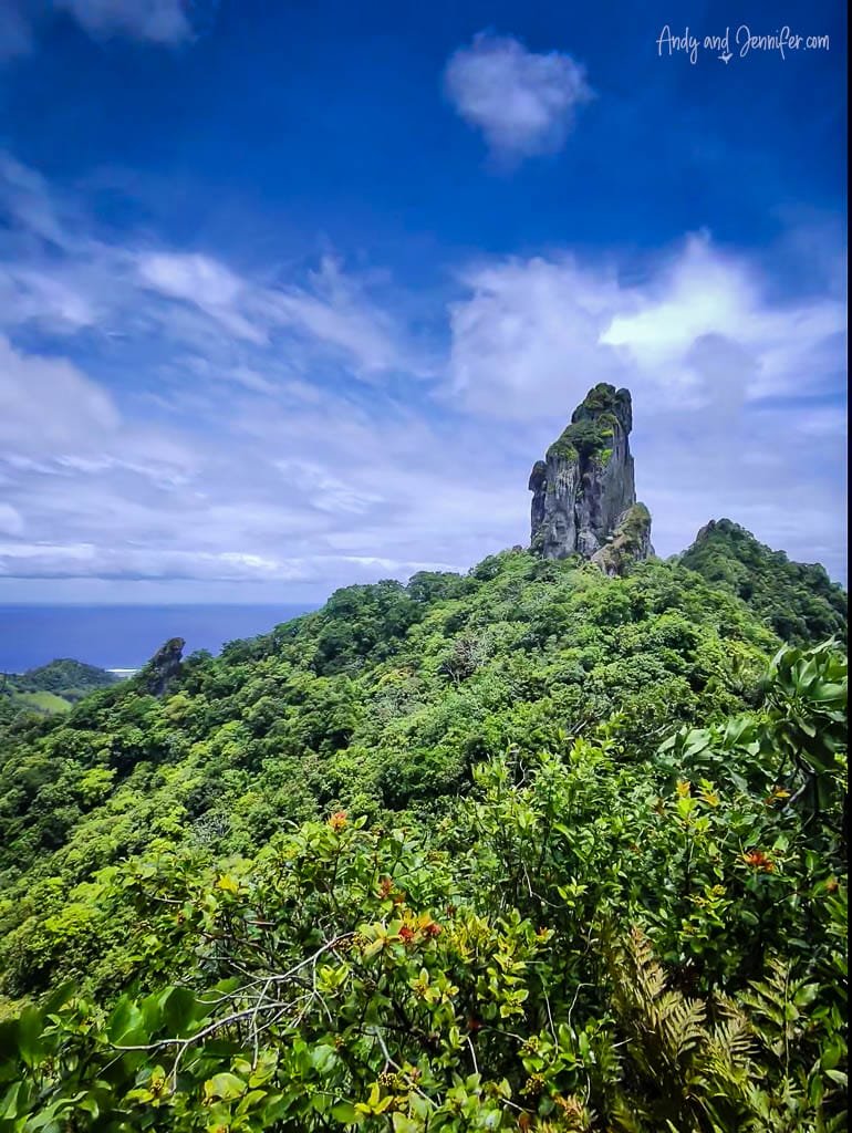 Towering rock peak amid vibrant forest and blue sky backdrop.