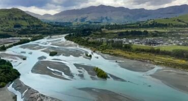 Winding river with sandbanks, green hills, and distant mountains under a cloudy sky.