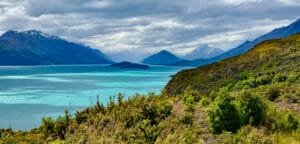 Turquoise lake surrounded by green hills and snow-capped mountains under cloudy skies