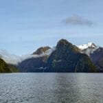 Serene fjord with forested cliffs, snow-capped peaks, and low-lying clouds