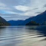 Calm reflective fjord surrounded by forested mountains under a partly cloudy sky.