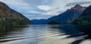 Calm reflective fjord surrounded by forested mountains under a partly cloudy sky.