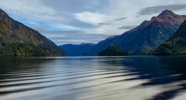 Calm reflective fjord surrounded by forested mountains under a partly cloudy sky.