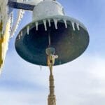Ship’s bell with hanging rope and icicles against blue sky.