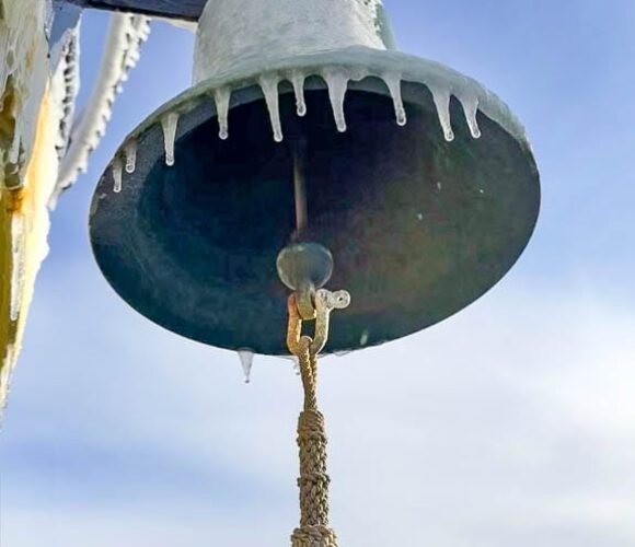 Ship’s bell with hanging rope and icicles against blue sky.