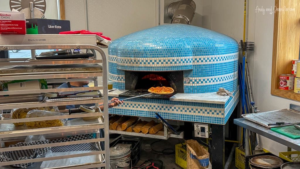 A chef uses a peel to remove a freshly baked pizza from a large blue-tiled wood-fired oven inside a pizzeria kitchen.