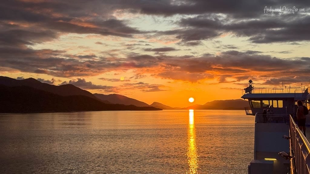 Sunset over calm water with mountains in the distance, viewed from a ferry deck.