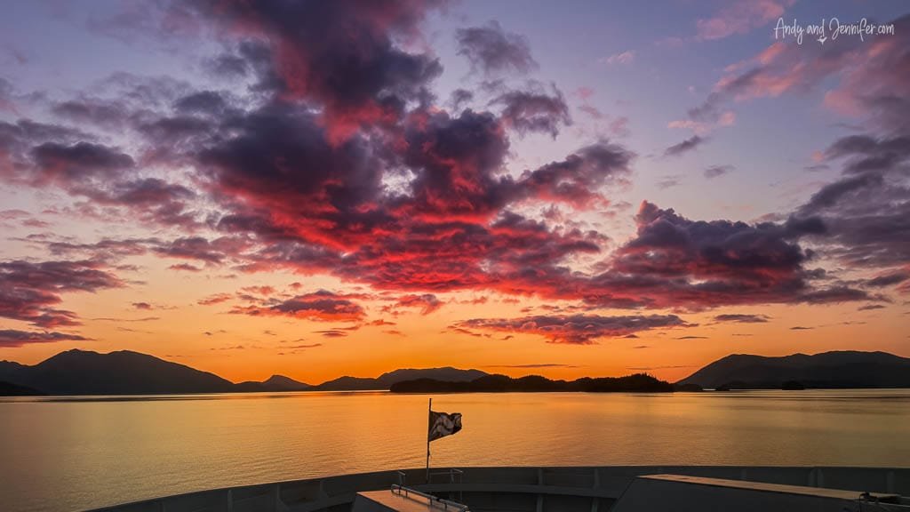 Vibrant pink and orange sunset sky over coastal mountains and ocean, seen from the front of a ferry.